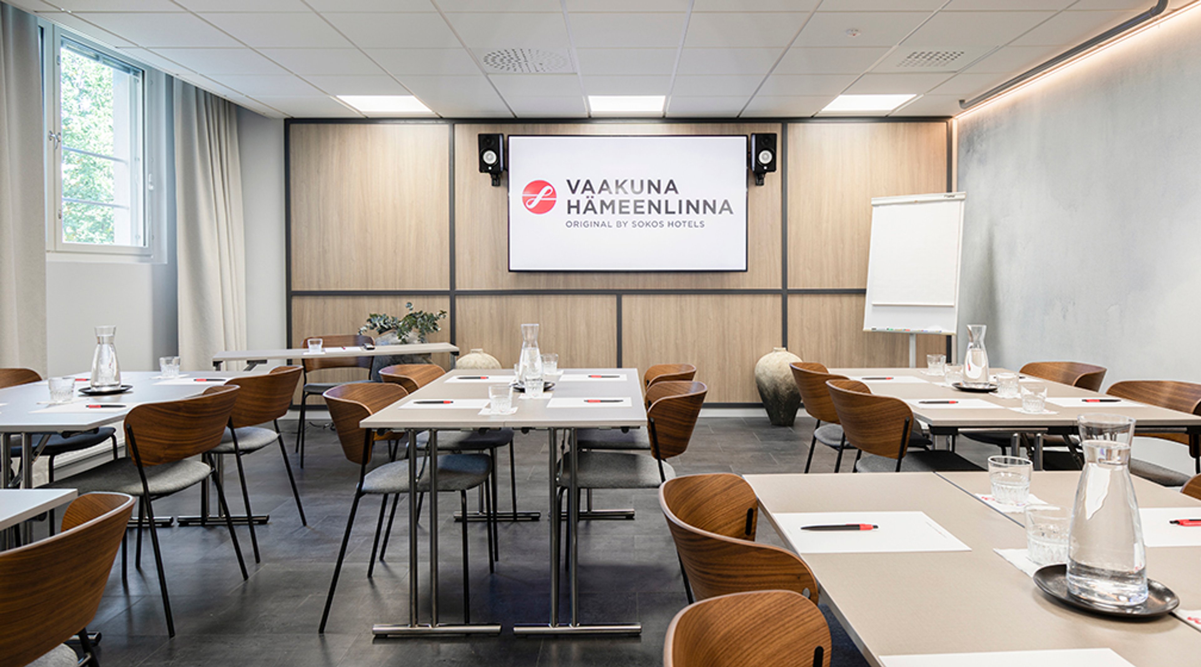 Bright meeting room with wooden chairs and white tables. Presentation screen at front, greenery visible through window, flipchart on the side.