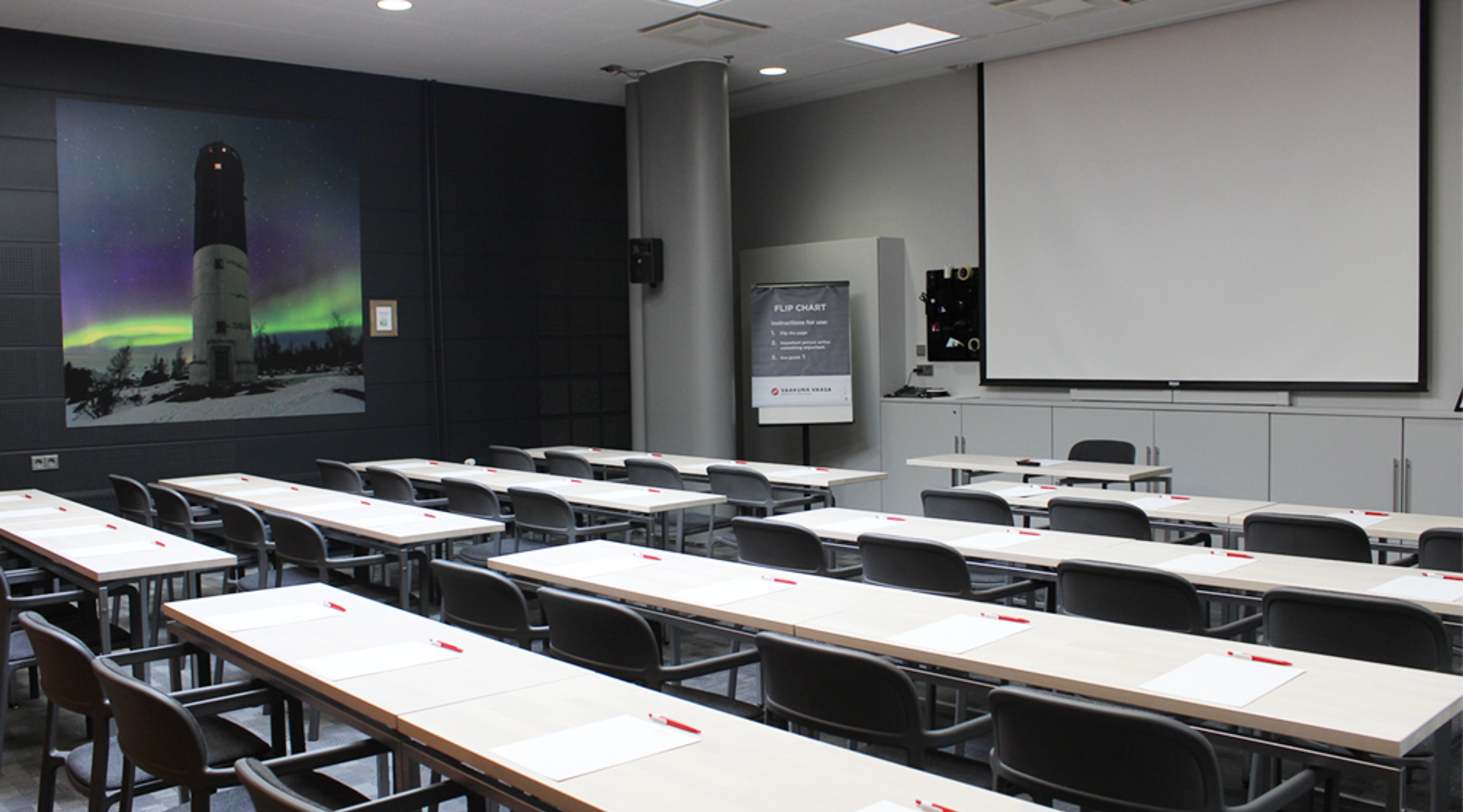 Classroom with long tables and chairs. Large image of a lighthouse under northern lights on the wall, projection screen at the front.