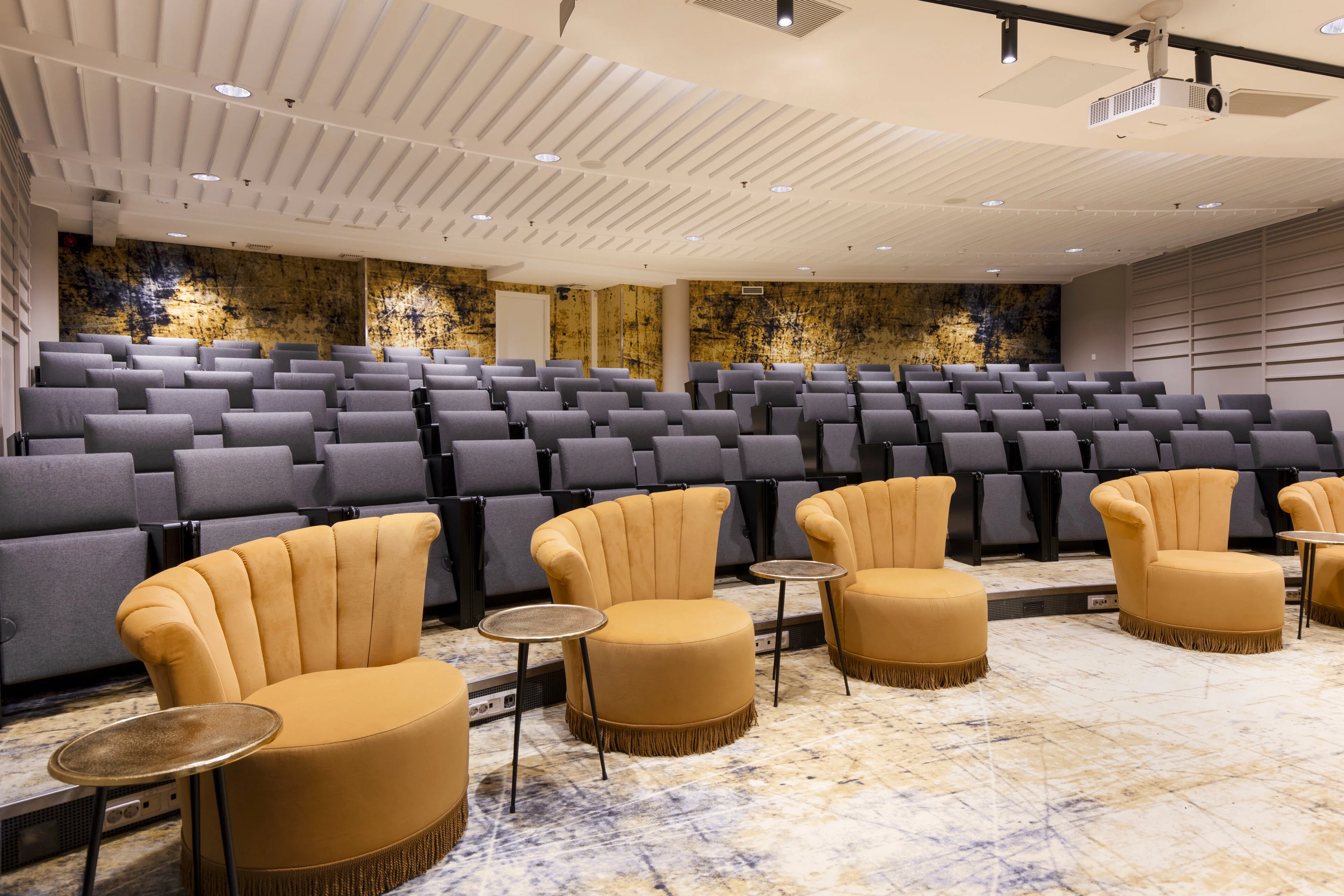 Moderni auditorio harmailla istuimilla, etualalla neljä keltaista nojatuolia ja pöydät. Kultainen taideseinä taustalla.