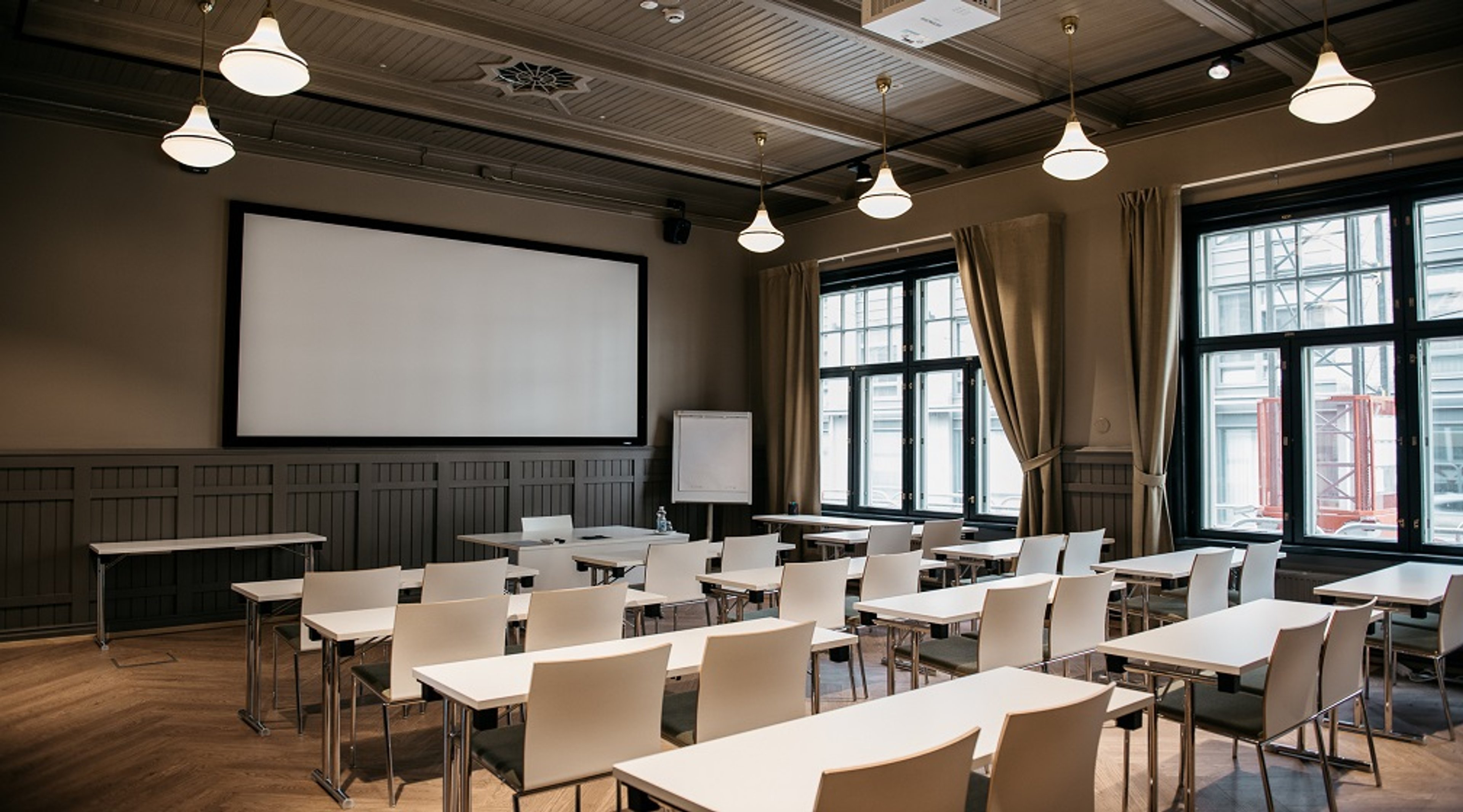 Classroom-style meeting room with white tables and chairs. Projection screen at front, large windows with beige curtains.