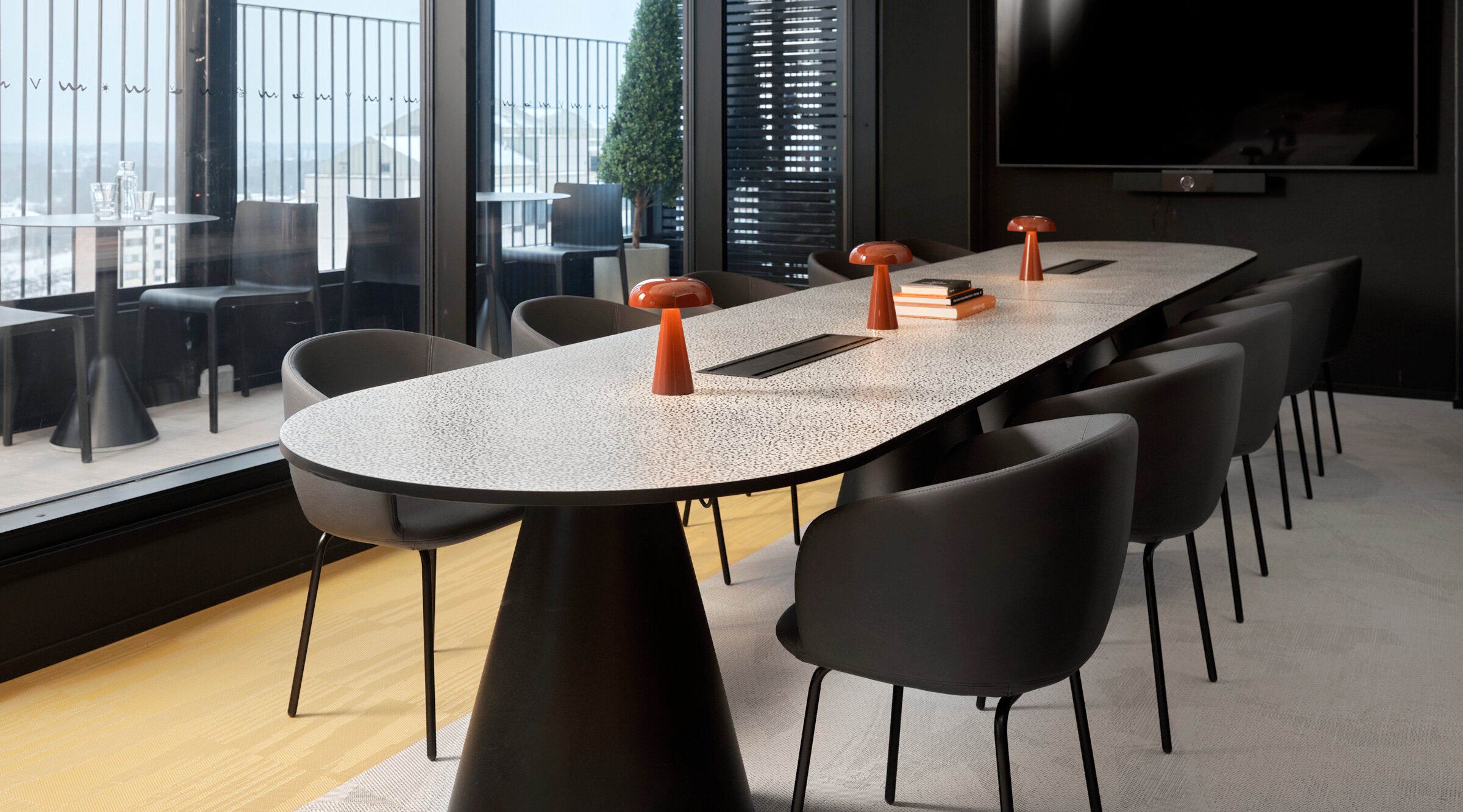 Modern conference room with white oval table and black chairs. Orange table lamps and large windows allowing natural light. Wooden floor and wall-mounted TV screen.