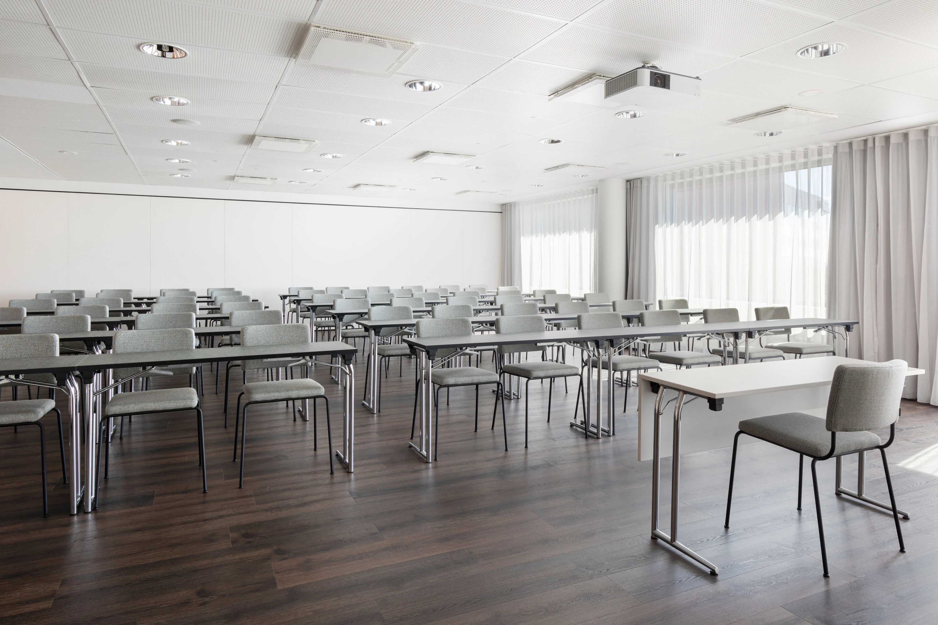 Bright conference room with gray tables and chairs arranged in rows. Wooden floor and ceiling-mounted projector. White walls and large windows with light curtains.