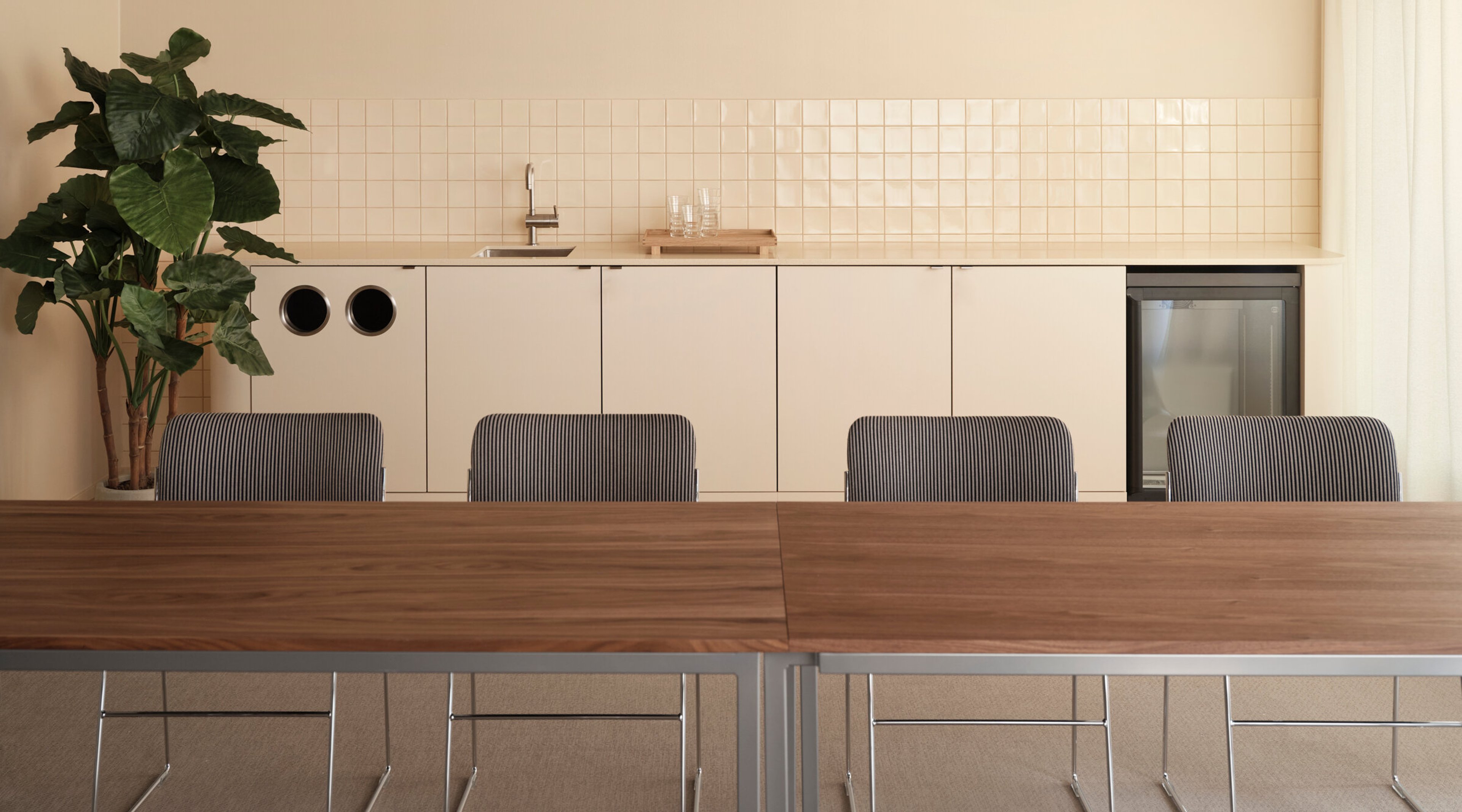 Minimalist meeting room with wooden table, striped chairs and beige kitchenette in background. Green plants add freshness to the space.