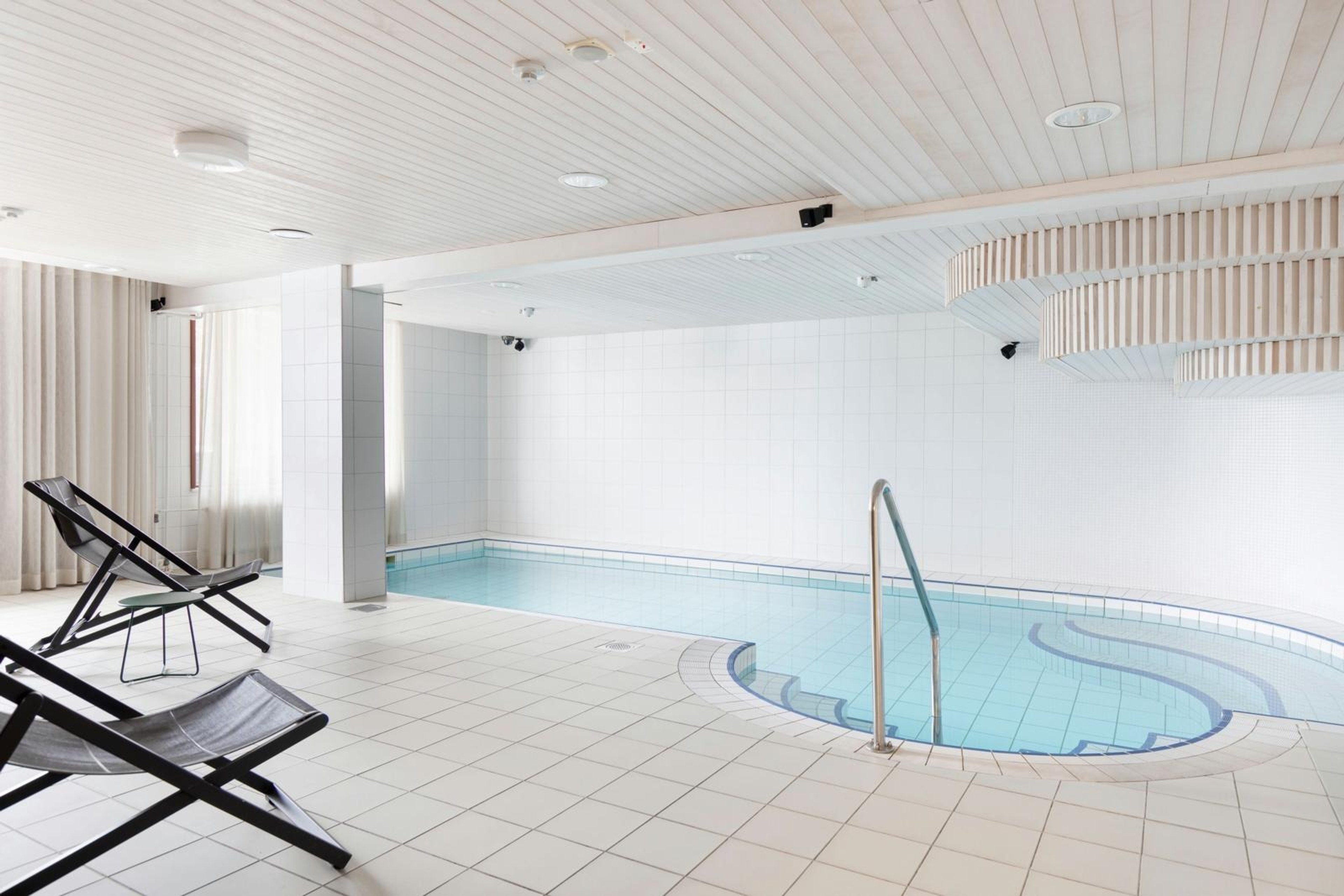 Ljus inomhuspool med vita kaklade väggar och golv. Metallräcke till poolen och några vilstolar intill.
